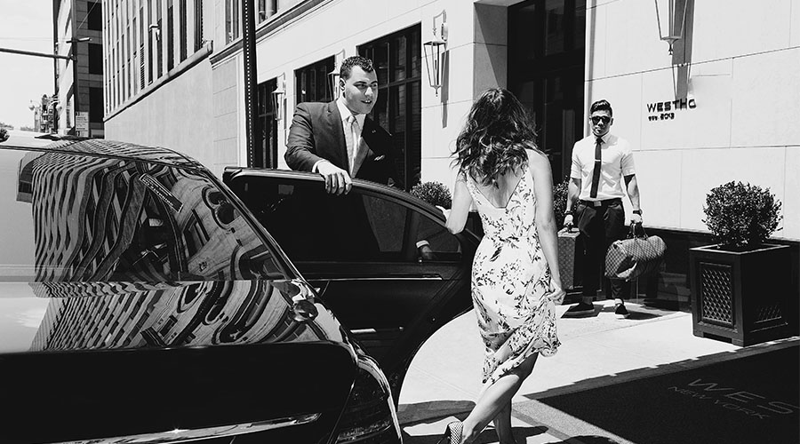 Image of a black car with the door opened by a hotel staff for a female guest. In front of the female guest is another hotel staff holding the guest luggage by the hotel entrance.